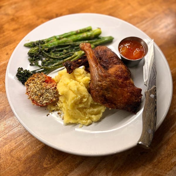 Crispy duck confit with mashed potatoes, asparagus, broccolini, and a breaded tomato on a white plate.