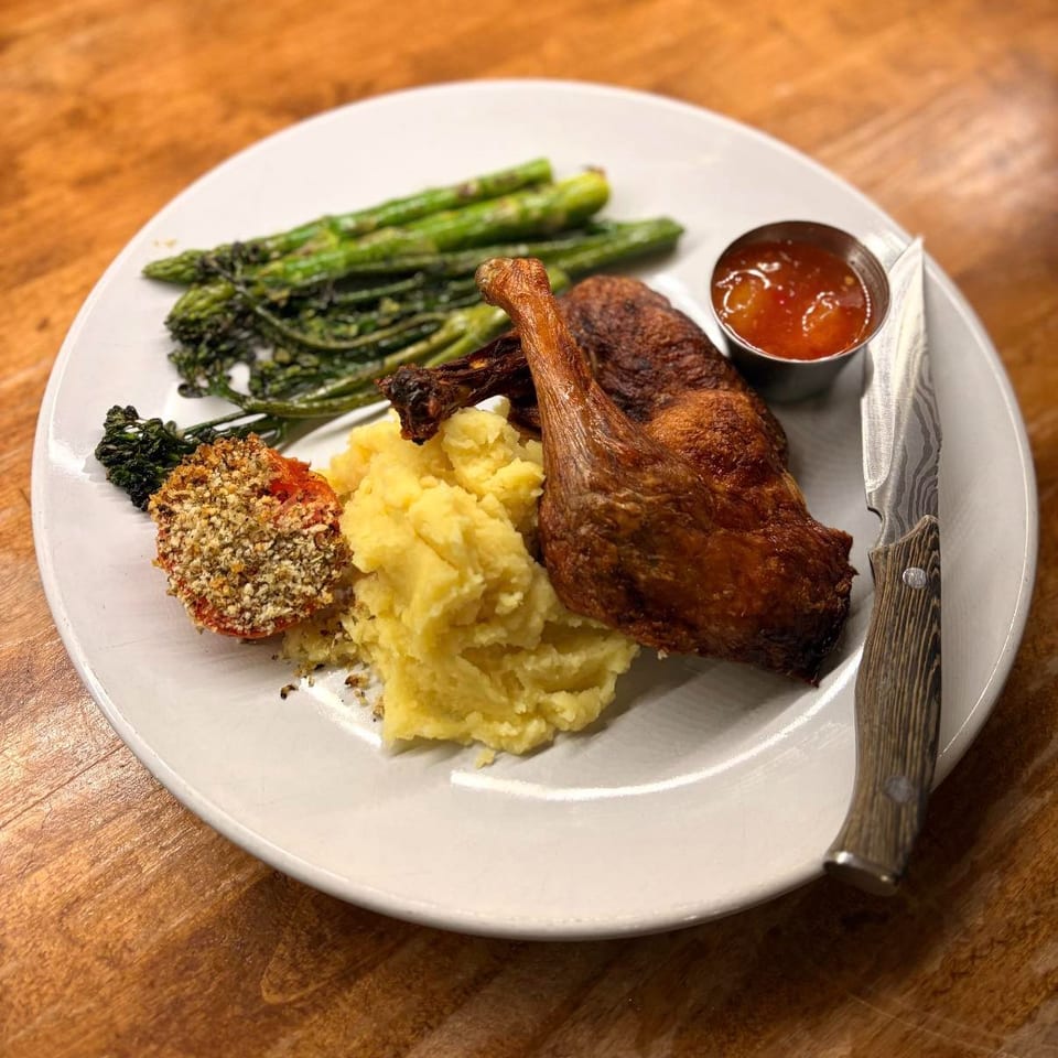 Crispy duck confit with mashed potatoes, asparagus, broccolini, and a breaded tomato on a white plate.