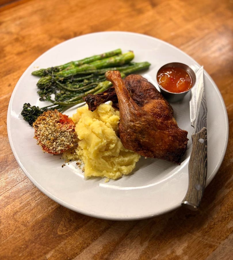Crispy duck confit with mashed potatoes, asparagus, broccolini, and a breaded tomato on a white plate.