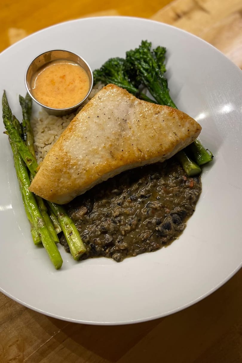 Pan-seared fish with rice, black beans, asparagus, broccolini, and a side of sauce.