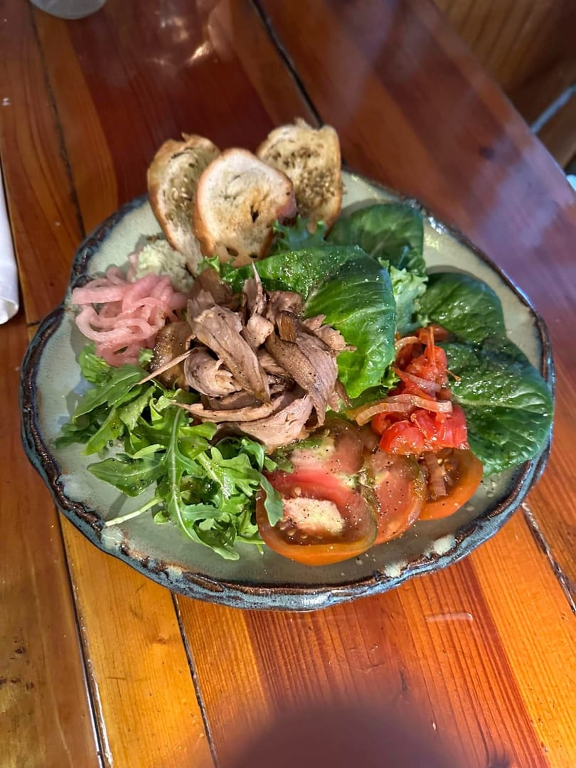 A vibrant salad with shredded meat, fresh greens, sliced tomatoes, pickled onions, and toasted bread.