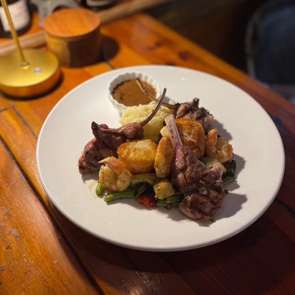 A white plate with lamb chops, shrimp, scallops, mashed potatoes, and vegetables on a wooden table.