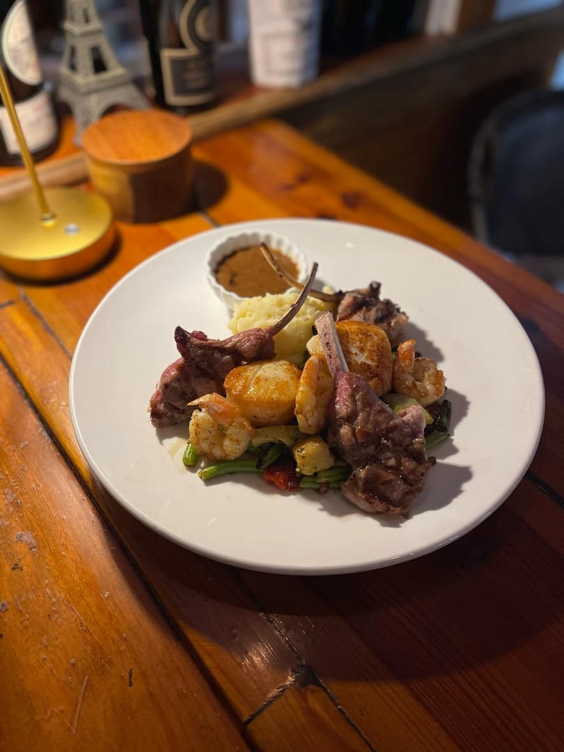 A white plate with lamb chops, shrimp, scallops, mashed potatoes, and vegetables on a wooden table.