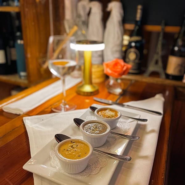 Three small bowls of soup on a white plate with spoons, a wine glass, and a table lamp.