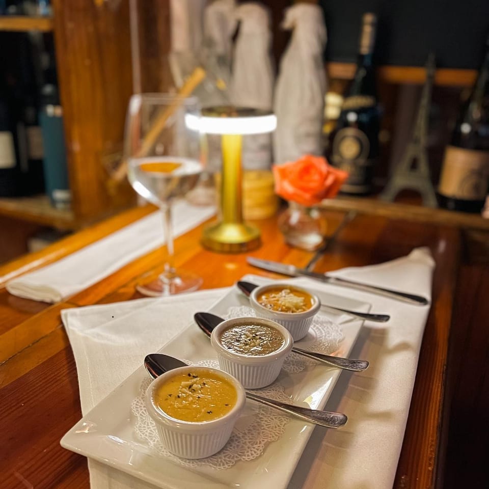 Three small bowls of soup on a white plate with spoons, a wine glass, and a table lamp.