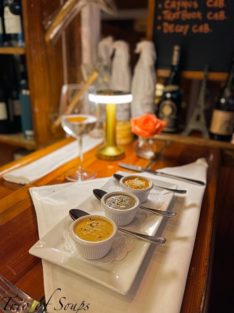 Three small bowls of soup on a white plate with spoons, a wine glass, and a table lamp.