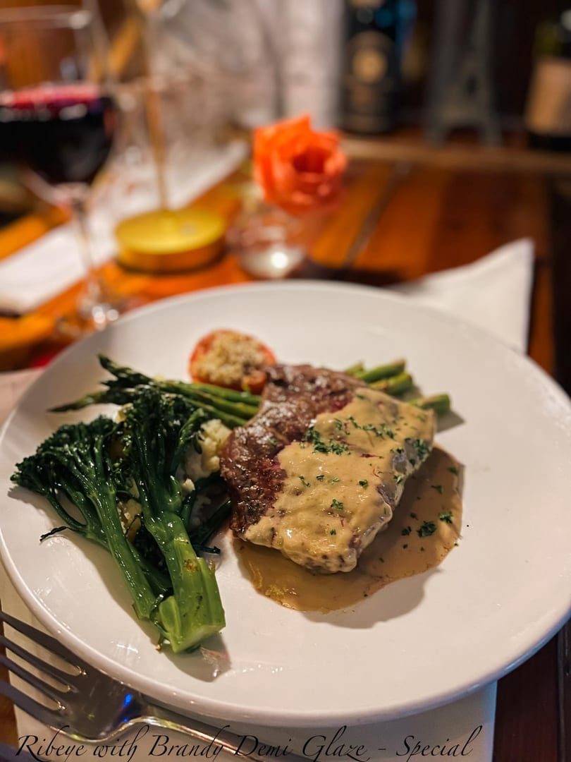 Ribeye steak with brandy demi-glaze, broccolini, asparagus, and a baked tomato on a white plate.