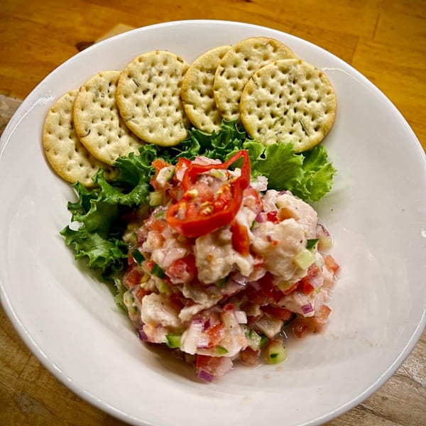 A white bowl of ceviche with red peppers, lettuce, and crackers on a wooden table.