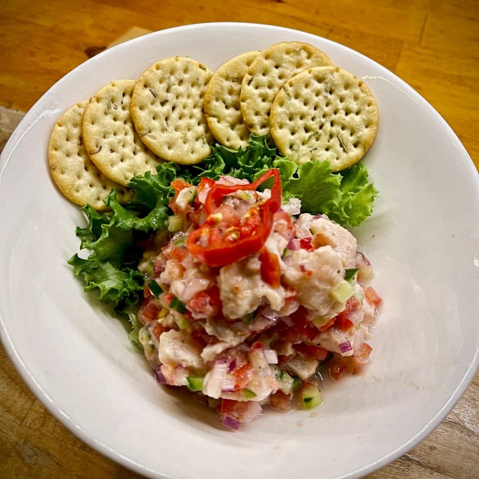 A white bowl of ceviche with red peppers, lettuce, and crackers on a wooden table.