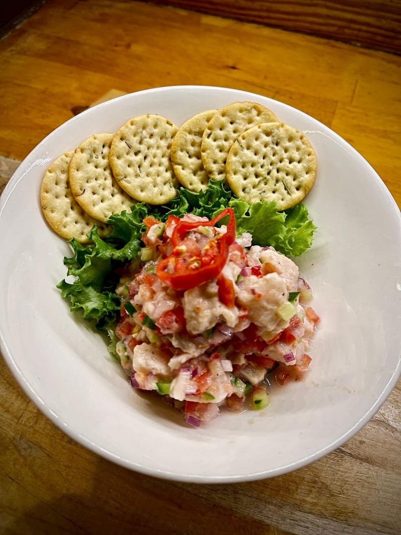 A white bowl of ceviche with red peppers, lettuce, and crackers on a wooden table.