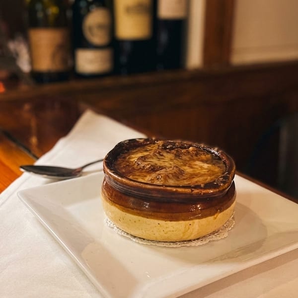 French onion soup in a crock with melted cheese, on a white plate with a rose and wine bottles.