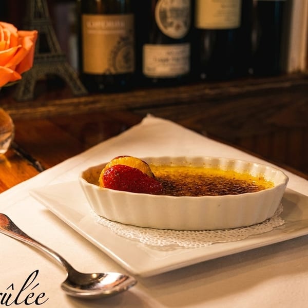 Crème brûlée with strawberry and orange garnish, spoon, and rose on a wooden table.